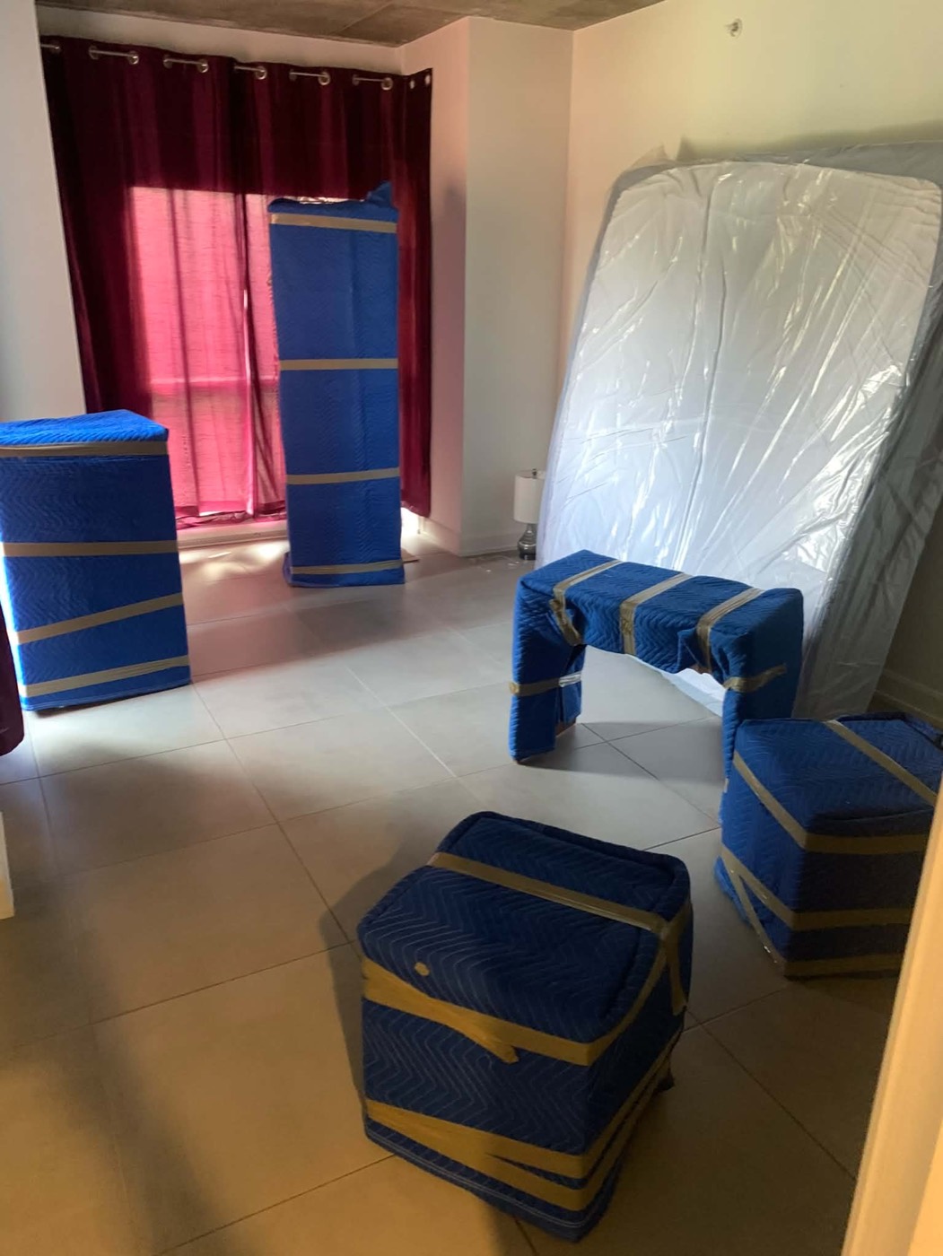 Dresser, cabinet and mattress wrapped in blue moving blankets and plastic in a bedroom